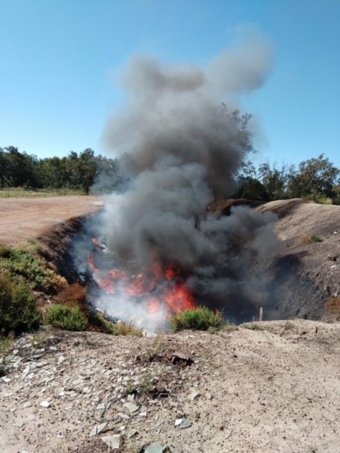 A pit filled with general waste is on fire. Flames extend above the top of the pit and thick black smoke billows from the pile.